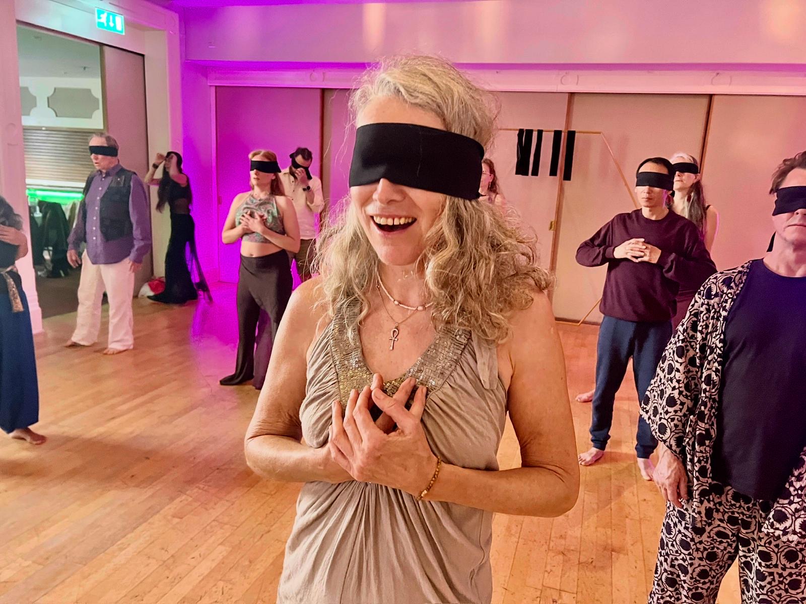 A woman smiling broadly while wearing a blindfold, hands resting at her heart, with other blindfolded participants behind her