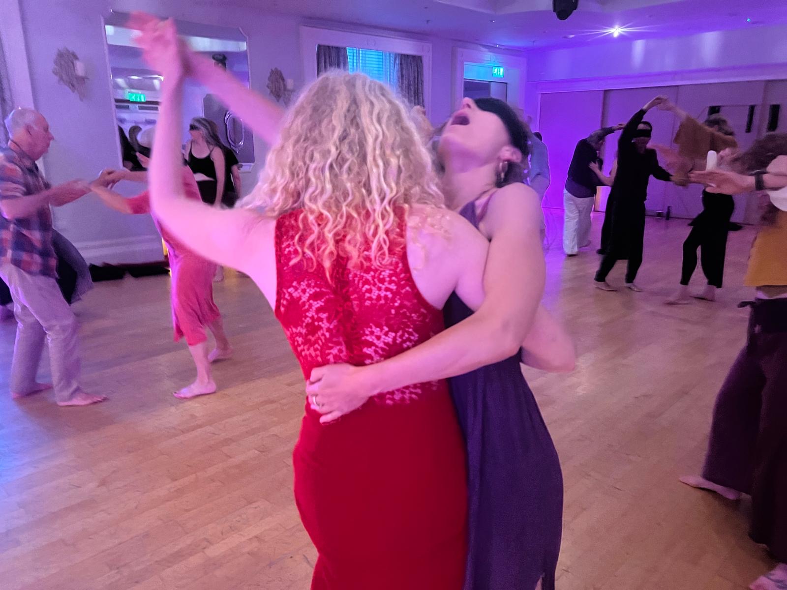 Dancers at our Devon gathering: two women dancing closely together, one in a red lace dress and one blindfolded in purple, as other pairs dance around them